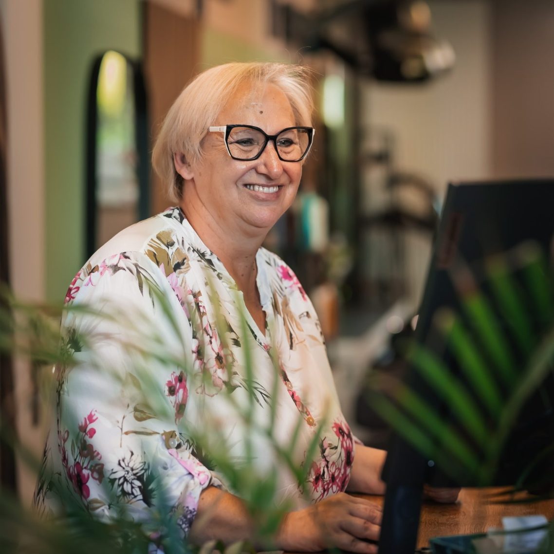 Frau mit grauen Haaren, Brille und schwarzer Kleidung vor einer Wand mit Scherenmotiven.