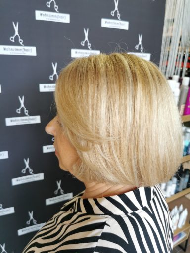 Eine Frau mit schulterlangem, blonden Haar in einem bunten Kleid steht vor einem Friseursalon.