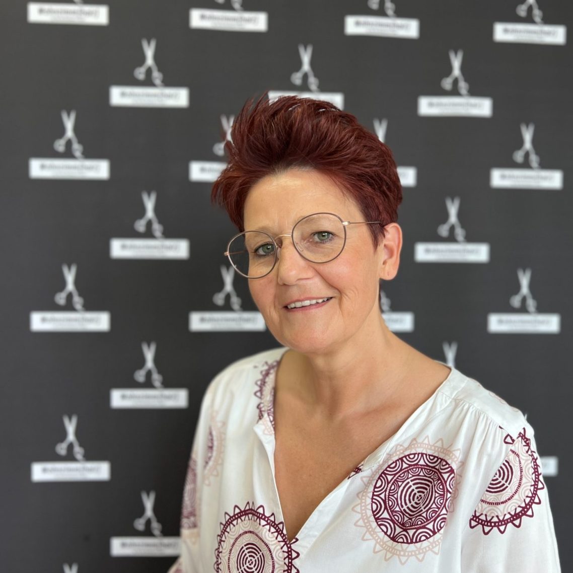 Frau mit kurzen, roten Haaren und Brille, trägt eine weiße Bluse mit Muster.