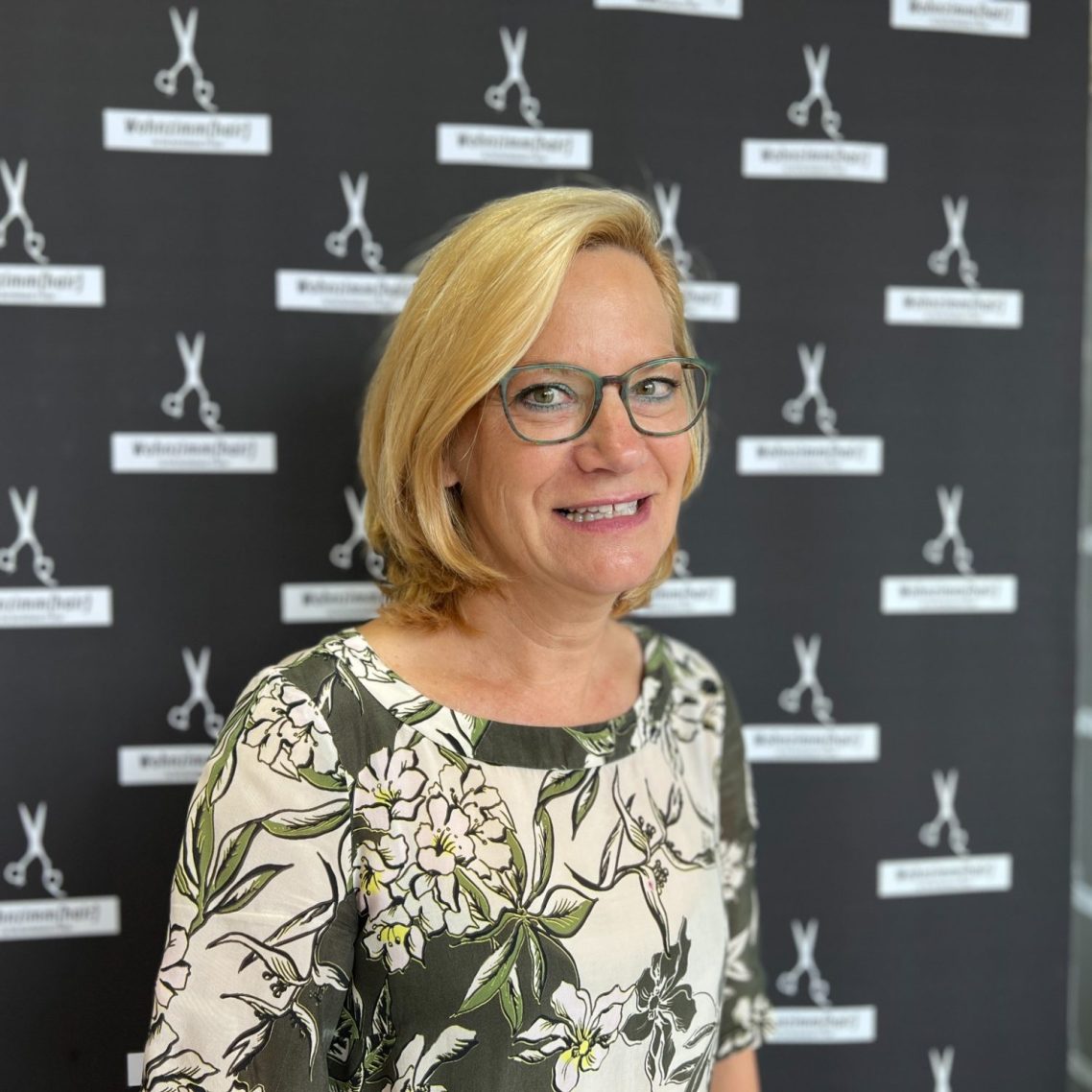 Eine lächelnde Frau mit blonden Haaren und Brille vor einem schwarzen Hintergrund.