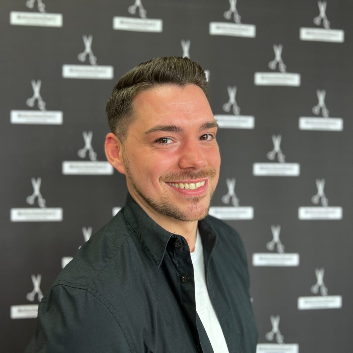 Lächelnder Mann in schwarzem Hemd vor schwarzem Hintergrund mit Logo.