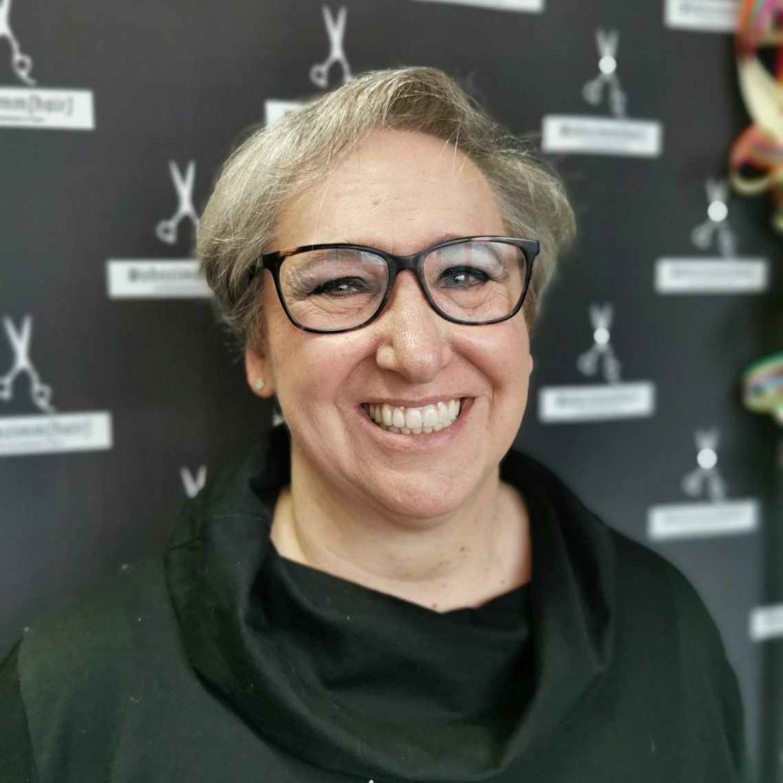 Frau mit grauen Haaren, Brille und schwarzer Kleidung vor einer Wand mit Scherenmotiven.