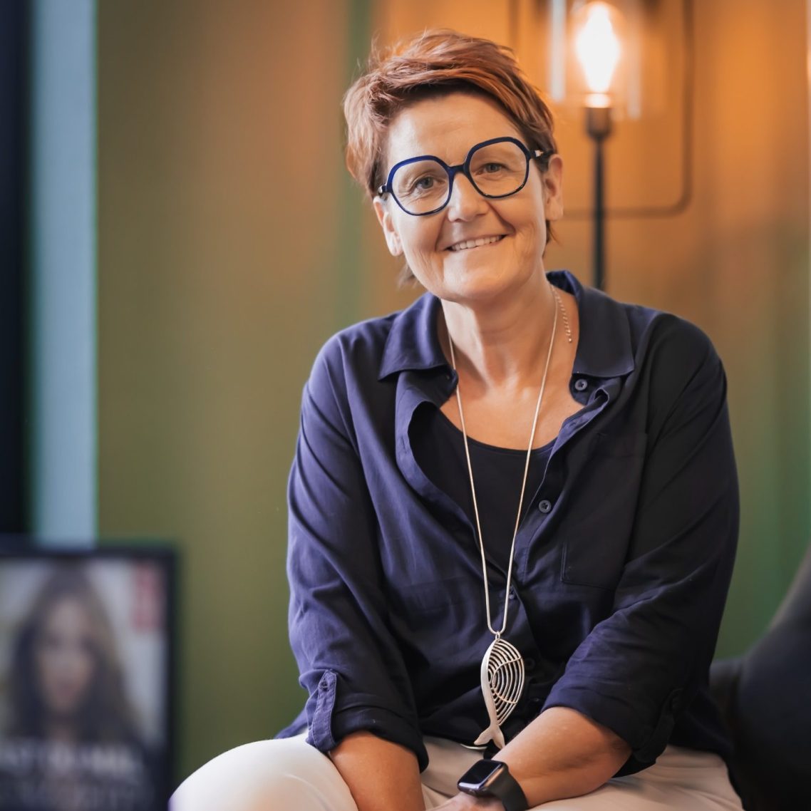 Frau mit kurzen, roten Haaren und Brille, trägt eine weiße Bluse mit Muster.